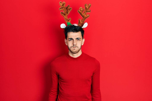 Jeune homme hispanique portant de jolies cornes de rennes de Noël gonflant les joues avec un drôle de visage. bouche gonflée d'air, expression folle.  - Photo, image