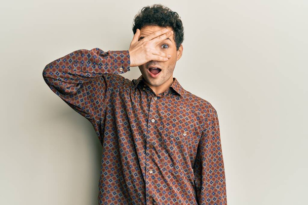 Handsome hispanic man wearing casual clothes peeking in shock covering face and eyes with hand, looking through fingers with embarrassed expression.  - Photo, Image