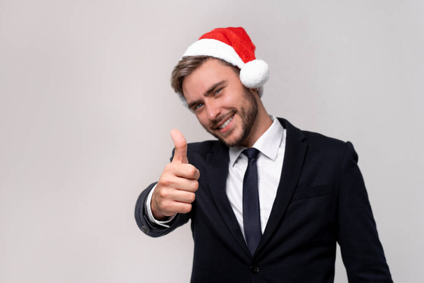 Young handsome caucasian guy in business suit and Santa hats on white background in studio smilie and showing thumbs up two hands. Close up portrait business person with Christmas mood Holiday banner - Zdjęcie, obraz