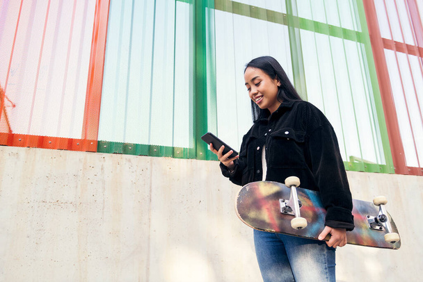 smiling young asian girl typing on the mobile phone standing outdoors with her skateboard in her hand - Foto, Imagen