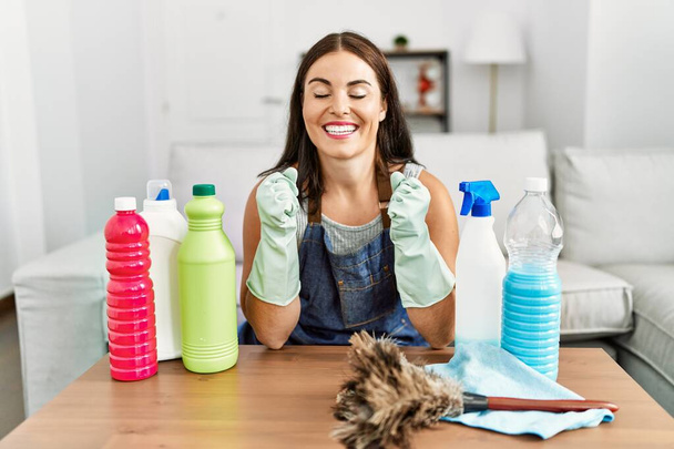 Young brunette woman wearing cleaner apron and gloves cleaning at home excited for success with arms raised and eyes closed celebrating victory smiling. winner concept.  - Photo, Image
