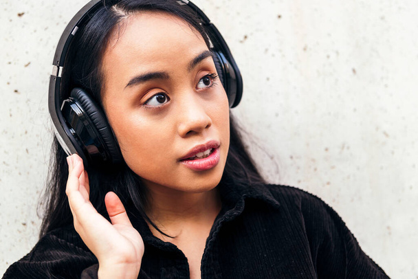 young asian woman enjoys listening to music with headphones outdoors leaning against a concrete wall - Photo, Image