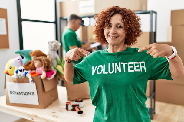 Középkorú nő visel önkéntes póló adományok állni néz magabiztos mosollyal az arcán, rámutatva önmagára ujjaival büszke és boldog.  - Fotó, kép