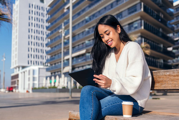asian young woman smiling sitting on a bench using her tablet next to a coffee - Photo, Image