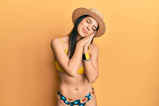 Młoda latynoska kobieta w bikini i letnim kapeluszu śpi zmęczona snem i pozuje dłońmi razem, uśmiechając się zamkniętymi oczami.  - Zdjęcie, obraz