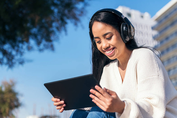 hymyilevä aasialainen nuori nainen kuulokkeet katsella videoita hänen tabletti istuu ulkona - Valokuva, kuva