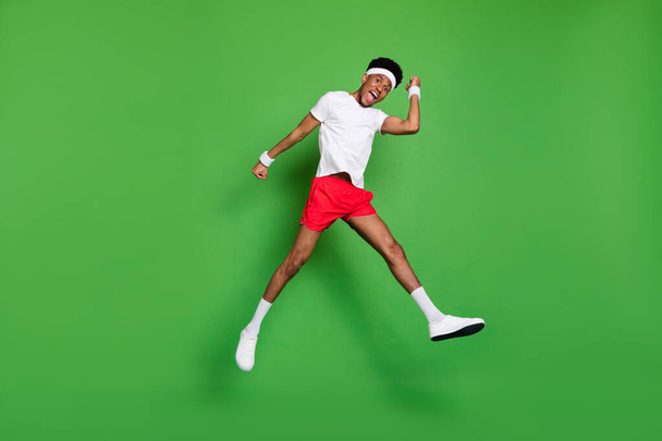 Full length photo of impressed excited dark skin man wear white t-shirt rising fists jumping high isolated green color background - Fotoğraf, Görsel