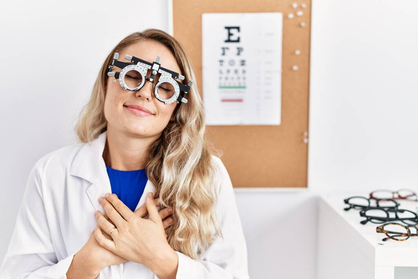 Klinikte optometri gözlüğü takan genç, güzel gözlüklü bir kadın elleri göğsünde, gözleri kapalı ve yüzünde minnet dolu bir jest ile gülümsüyor. sağlık kavramı.  - Fotoğraf, Görsel