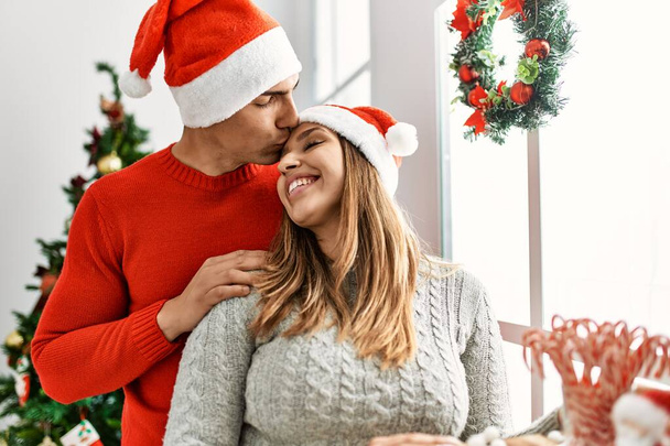 Młoda para uśmiechnięta szczęśliwa w świątecznym kapeluszu. Stojąc z uśmiechem na twarzy opierając się o okno w domu. - Zdjęcie, obraz