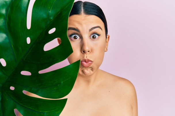 Όμορφη μελαχρινή γυναίκα κρατώντας πράσινο φύλλο φυτών κοντά στο όμορφο πρόσωπο puffing μάγουλα με αστείο πρόσωπο. στόμα φουσκωμένο με αέρα, που πιάνει αέρα.  - Φωτογραφία, εικόνα