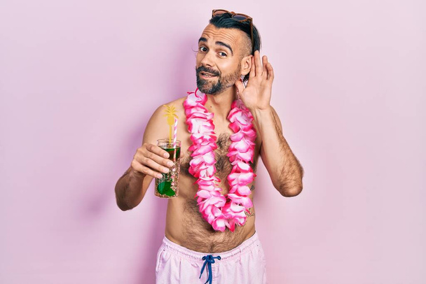Young hispanic man wearing swimsuit and hawaiian lei drinking tropical cocktail smiling with hand over ear listening an hearing to rumor or gossip. deafness concept.  - Photo, Image