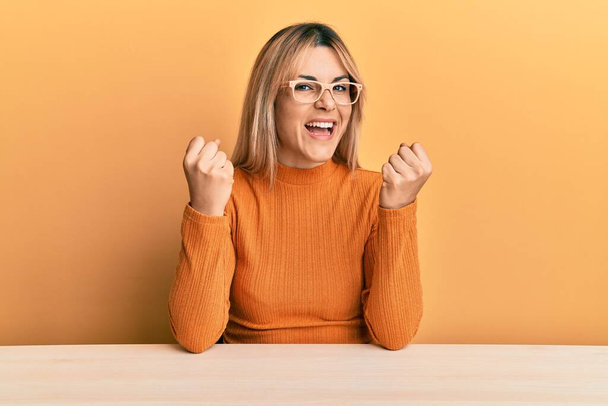 Young caucasian woman wearing casual clothes and glasses sitting on the table screaming proud, celebrating victory and success very excited with raised arms  - Photo, Image