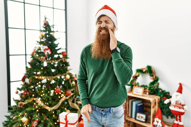 Rothaariger Mann mit langem Bart, der Weihnachtsmütze am Weihnachtsbaum trägt und den Mund mit schmerzverzerrter Hand berührt, weil Zahnschmerzen oder Zahnkrankheiten an den Zähnen auftreten. Zahnarzt  - Foto, Bild