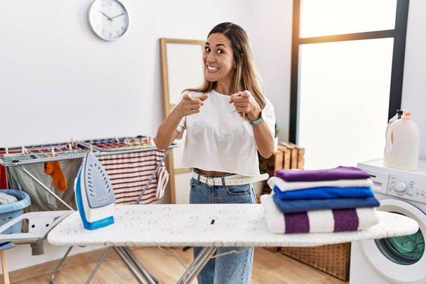 Молода іспаномовна жінка прасує одяг у пральні, вказуючи пальці на камеру з щасливим і смішним обличчям. хороша енергія і вібрує
.  - Фото, зображення