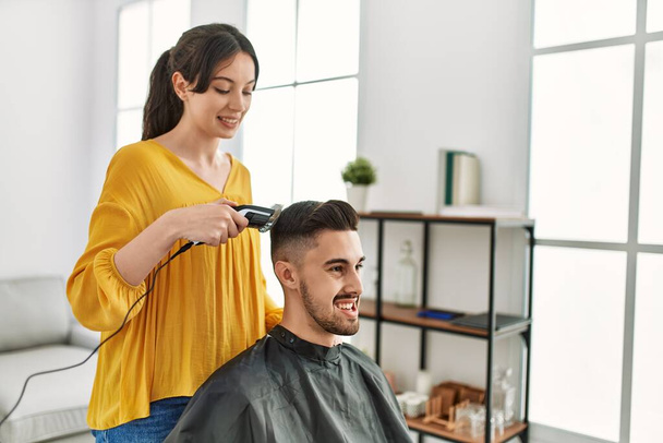 Młoda Latynoska fryzjerka obcinająca włosy mężczyźnie za pomocą maszynki do golenia elektrycznego w centrum urody. - Zdjęcie, obraz