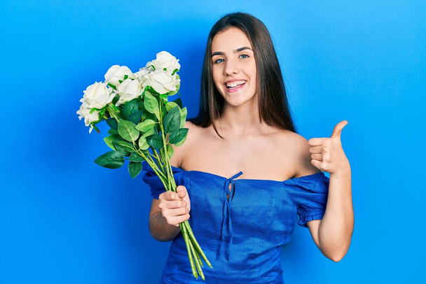 Young brunette teenager holding bouquet of white flowers smiling happy and positive, thumb up doing excellent and approval sign  - Photo, Image