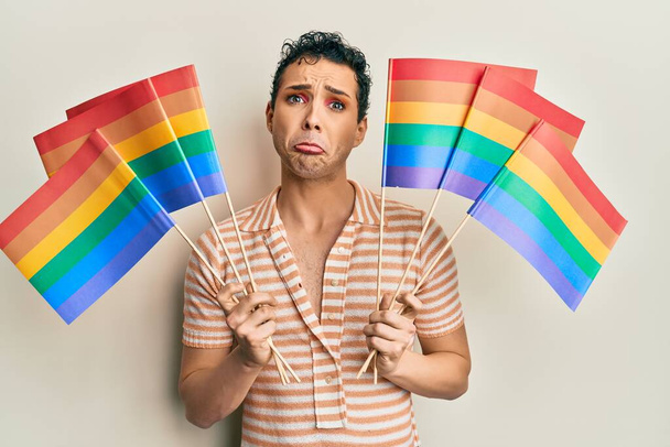 Knappe man draagt make-up houden regenboog lgbtq vlaggen depressief en zorgen te maken voor ellende, huilen boos en bang. trieste uitdrukking.  - Foto, afbeelding