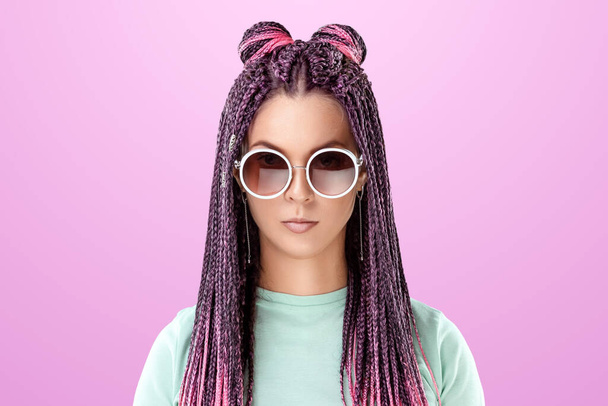 Beautiful girl with a hairstyle of pink braids in turquoise clothes and sunglasses posing on a pink background in the studio. The concept is modern style, individuality, inclusiveness, creativity - Фото, зображення