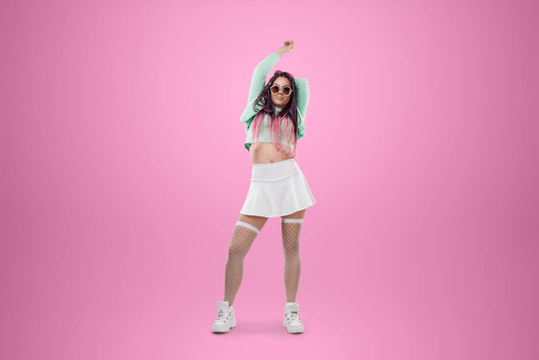 Millennial girl with pink braids hairstyle in turquoise clothes and sunglasses posing on a pink background. Concept for modern style, inclusiveness, individuality, generation Z, copy space - Photo, image