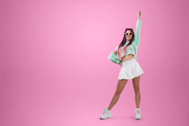 Millennial girl with pink braids hairstyle in turquoise clothes and sunglasses posing on a pink background. Concept for modern style, inclusiveness, individuality, generation Z, copy space - Fotó, kép