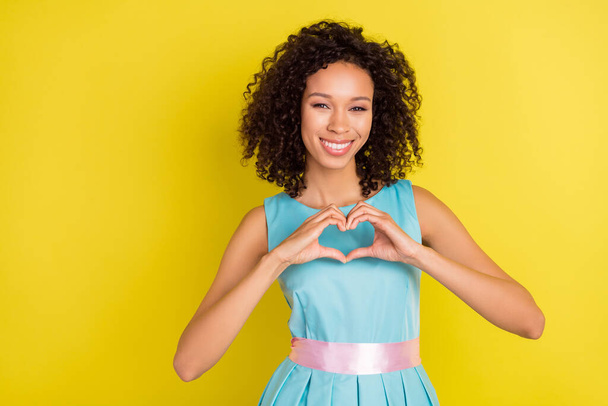 Photo of sweet pretty dark skin woman wear blue outfit showing arms heart empty space smiling isolated yellow color background - Фото, изображение