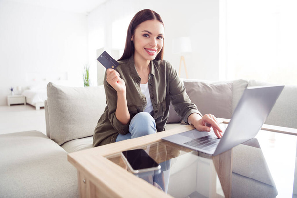 Photo of sweet adorable young lady wear green shirt sitting sofa typing modern device holding credit card indoors room home house - 写真・画像