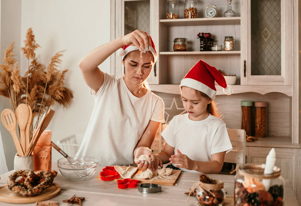 мама и дочь в рождественских шляпах учатся готовить печенье на кухне - Фото, изображение
