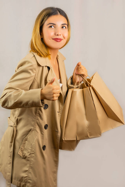 Portrait of a young Asian woman in a trench coat holding paper craft gift bags and showing what she likes. Concept: shopping black friday  - Foto, imagen