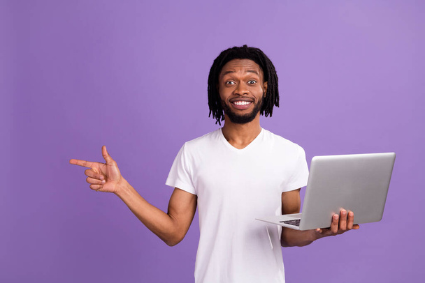 Photo of amazed young dark skin brown dreadlocks haired man point empty space hold laptop isolated on violet color background - Photo, Image