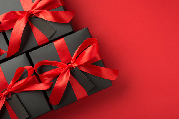 Top view photo of black gift boxes with red ribbon bow and tags on isolated red background with blank space - Zdjęcie, obraz