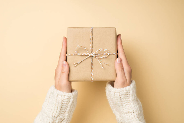 First person top view photo of girl hands in white knitted pullover holding craft paper giftbox with twine bow on isolated beige background - Photo, Image