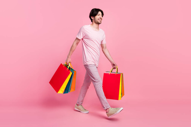 Full body profile side photo of young man happy positive smile go walk hold shopping bags isolated over pink color background - 写真・画像