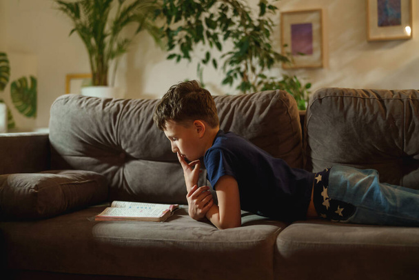 An attentive boy is reading a book lying on the couch at home. The student is passionate about reading. The child prepares homework, does homework lying down. - Foto, afbeelding