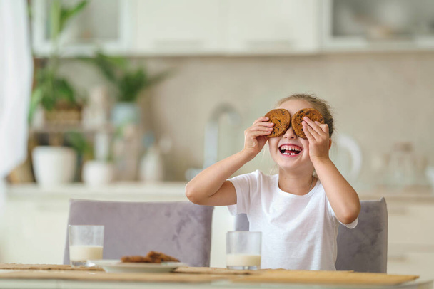 little girl is sitting at table during breakfast, having fun and playing with oatmeal cookies in her hands. child enjoys healthy natural products, having in kitchen. Proper nutrition for children. - 写真・画像