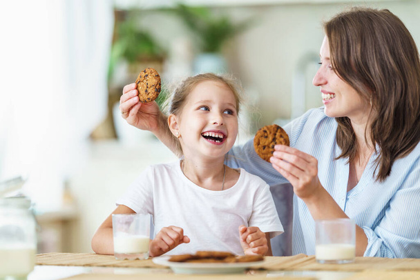 happy loving family, mom and daughter, playing sitting at table and having breakfast at home in morning. woman and girl eat oatmeal cookies and drink cow's milk, and have nice time together in kitchen - Photo, image