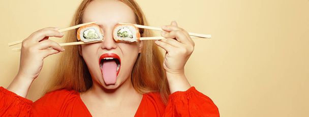 Piękna dziewczyna jedząca sushi na białym talerzu. Kobieta w czerwonej sukience i czerwonych ustach je sushi z łososiem z Filadelfii. Modelka robi minę i dobrze się bawi zamykając oczy rolką. - Zdjęcie, obraz
