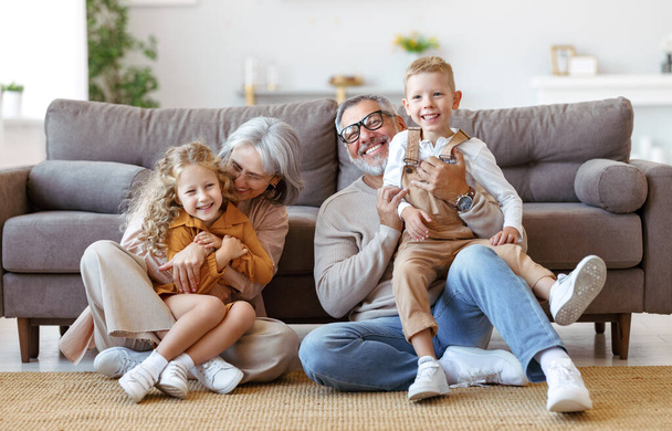 Happy rodina prarodiče a dvě malé děti objímající a usmívající se na kameru, zatímco sedí na podlaze - Fotografie, Obrázek
