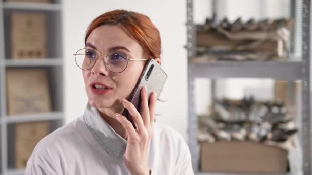 online werk, jonge vrouw met bril praat op mobiele telefoon met de klant en neemt een bestelling voor gedroogde vis terwijl zitten in het magazijn op de achtergrond van planken - Video