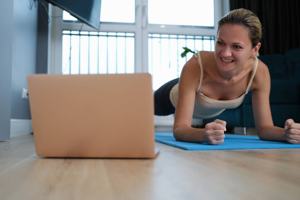Young woman standing in plank on floor in front of laptop - 写真・画像