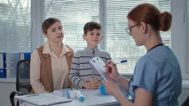 gabinet szpitalny, lekarka pisze do schowka i mierzy temperaturę chłopca z bezdotykowym termometrem - Materiał filmowy, wideo