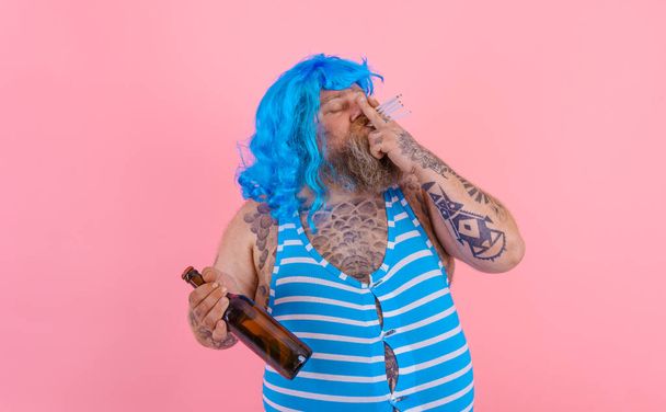 Fat man with beard and wig smokes cigarettes and drinks beer - Photo, Image