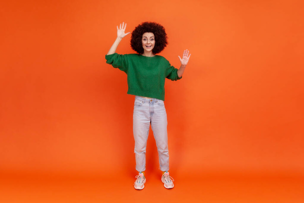 Full length portrait of woman with Afro hairstyle wearing green casual style sweater standing with raised arms, waving hello, glad to meet you. Indoor studio shot isolated on orange background. - 写真・画像
