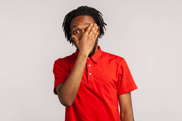 Portrait of young bearded man with dreadlocks wearing red casual style T-shirt, spying through hole in fingers closing eyes with palm, peeking. Indoor studio shot isolated on gray background - Фото, изображение