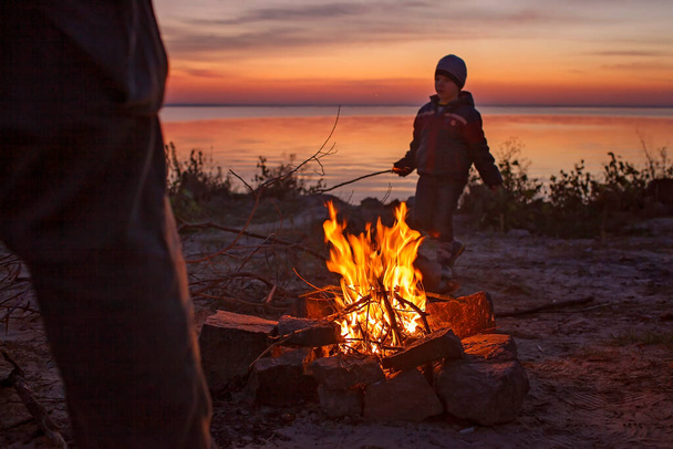 Dzieci siedzą przy ogniu na jesiennym brzegu morza po zachodzie słońca, rozgrzewają ręce, bawią się razem, na zewnątrz - Zdjęcie, obraz