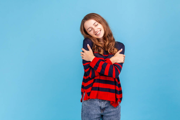 Portrait of lovely female in striped casual style sweater, standing hugging herself and feeling good, appreciating her appearance, self esteem concept. Indoor studio shot isolated on blue background. - Photo, Image