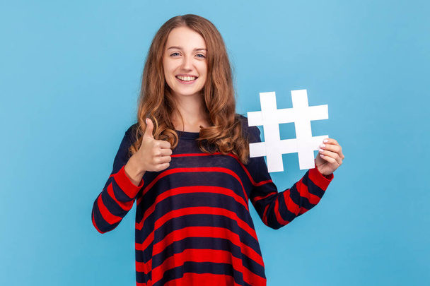 Happy woman wearing striped casual style sweater, showing hashtag and thumb up, promoting viral topic in social network, tagging blog trends. Indoor studio shot isolated on blue background. - Photo, Image