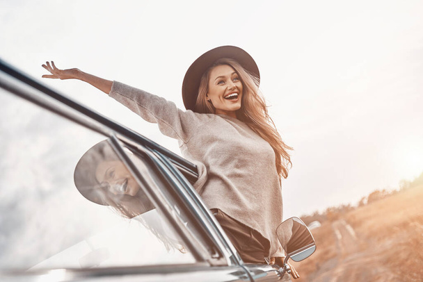 Beautiful young woman in hat keeping arms outstretched and looking happy while enjoying road trip in minivan - Photo, Image
