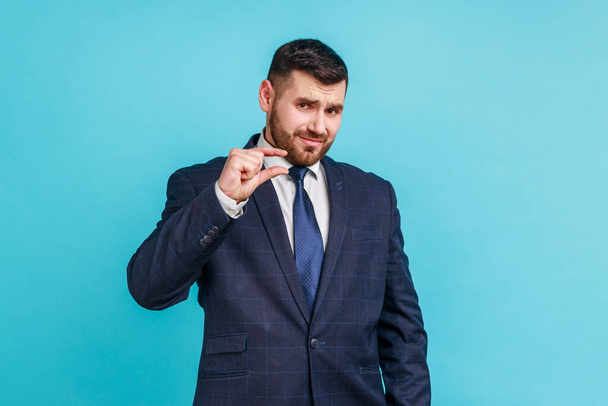 Handsome bearded businessman wearing official style suit showing a little bit gesture, small amount or low scale sign, few centimeters. Indoor studio shot isolated on blue background. - Fotoğraf, Görsel