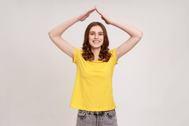 I'm in safety. Portrait of happy teenager girl with wavy hair in yellow t-shirt standing and raising hands, showing roof gesture, dreaming of house. Indoor studio shot isolated on gray background. - Photo, image