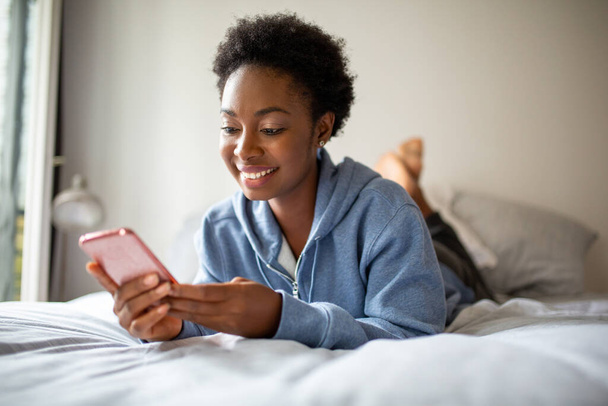 Portrait smiling young african american woman lying on bed looking at mobile phone - Φωτογραφία, εικόνα
